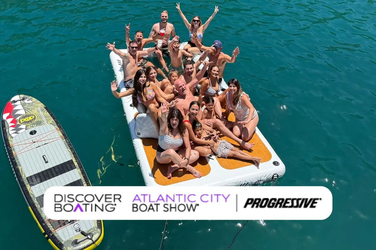 Large group of adults and children sitting together on an inflatable dock platform in clear blue ocean water, with a paddle board floating beside them.