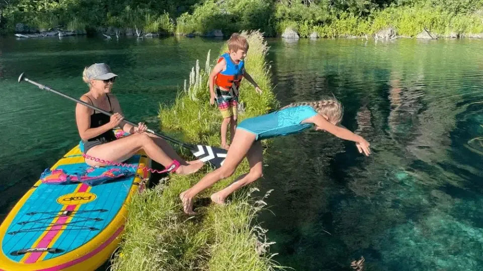 Family enjoying inflatable paddle board on clear lake with kids jumping into water