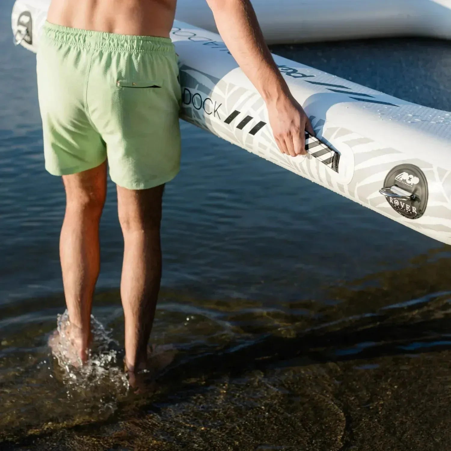 Close-up of a man carrying an Aqua Dock inflatable platform by its reinforced side handle while standing at the water’s edge.