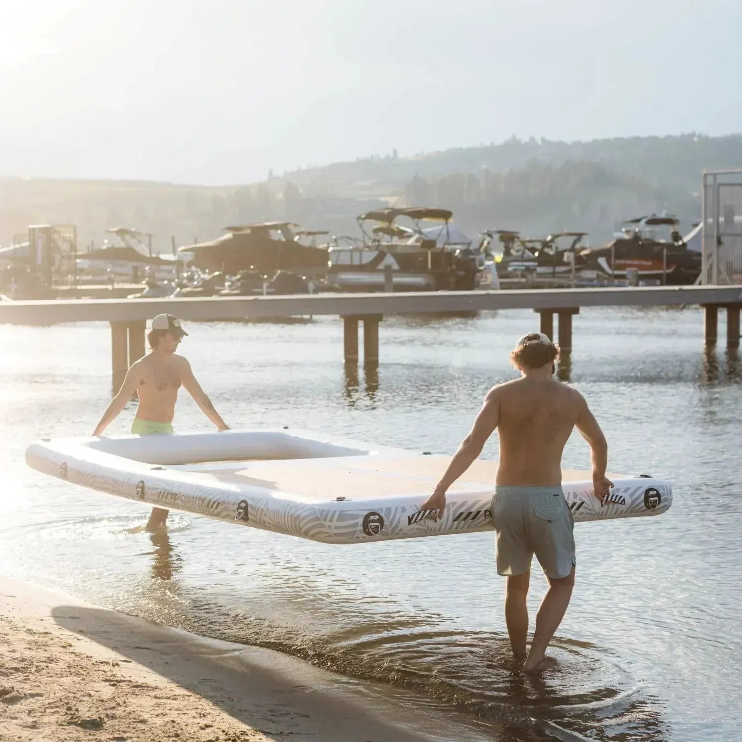 Two men carrying an Aqua Dock inflatable platform from the beach into the water at a marina, preparing to launch it for use.