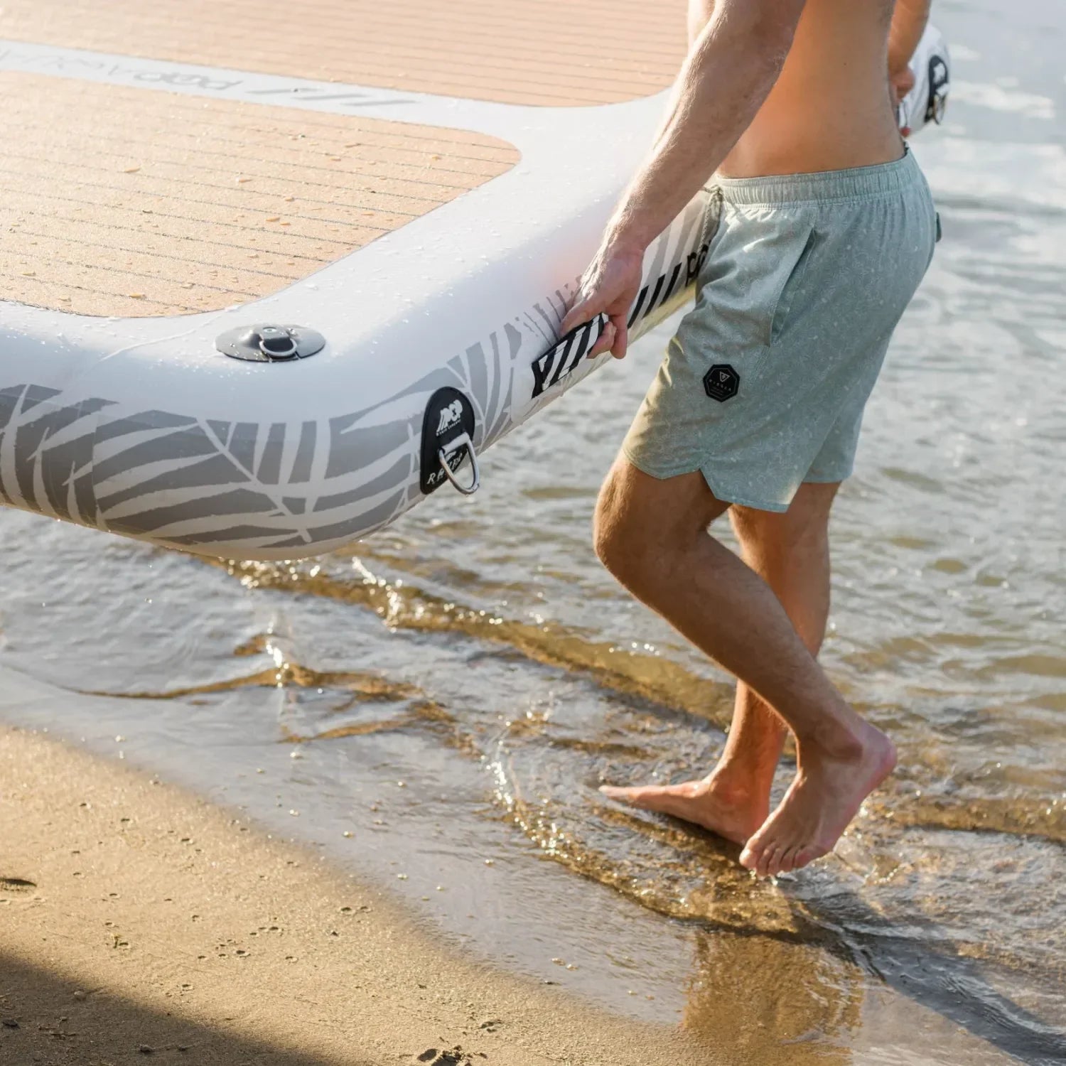 Man carrying an Aqua Dock inflatable platform from the shoreline, showing its durable handles, reinforced PVC construction, and wood-style traction pad