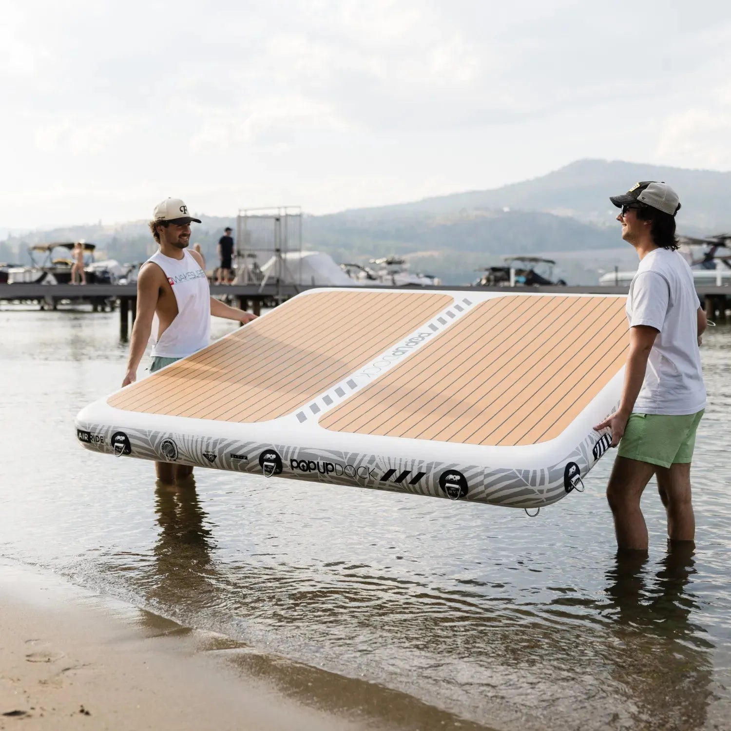 Two friends carrying a POPUP Dock inflatable platform at the shoreline, getting ready to launch it onto the water with boats and docks in the background.