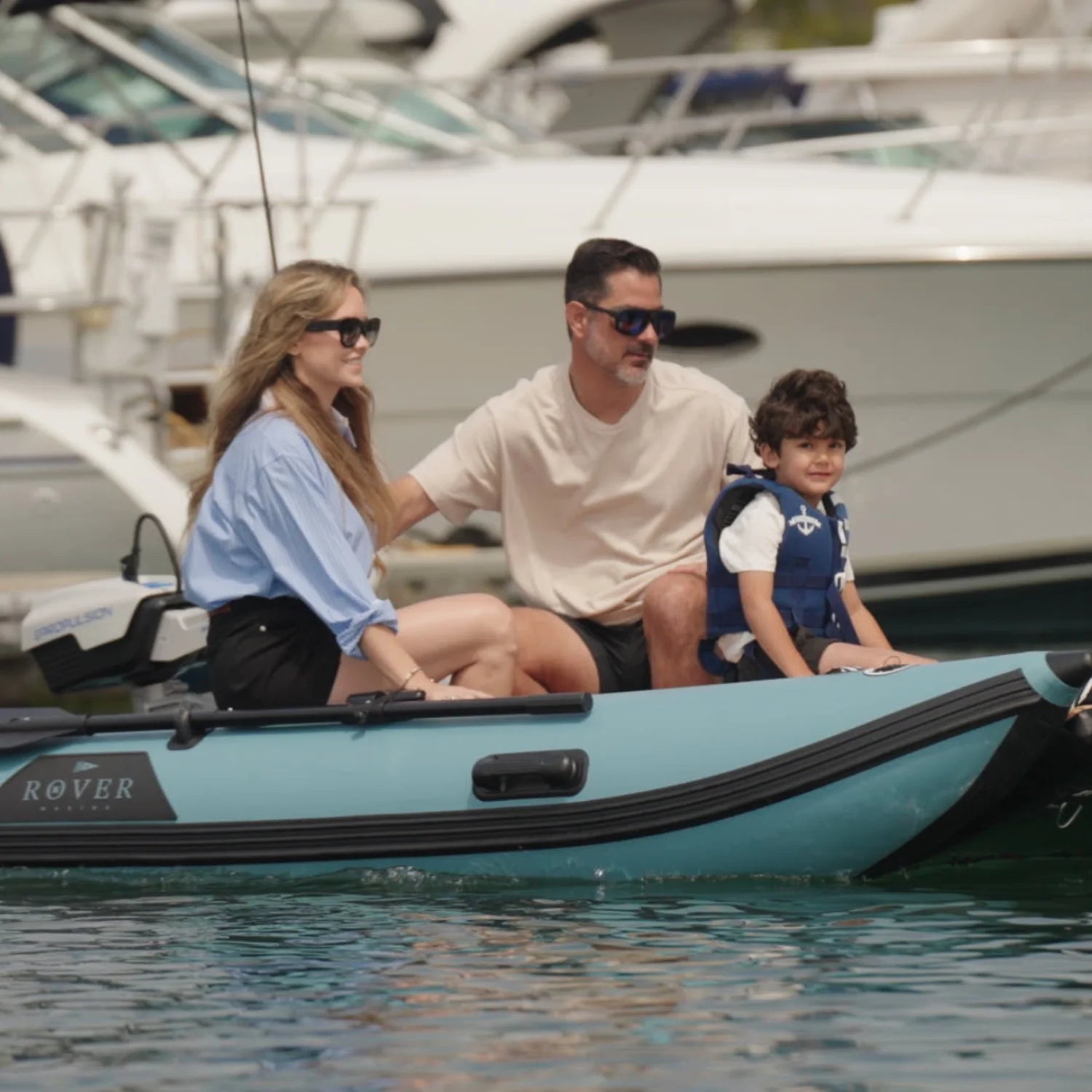 Family of three on a small boat in a marina with yachts in the background