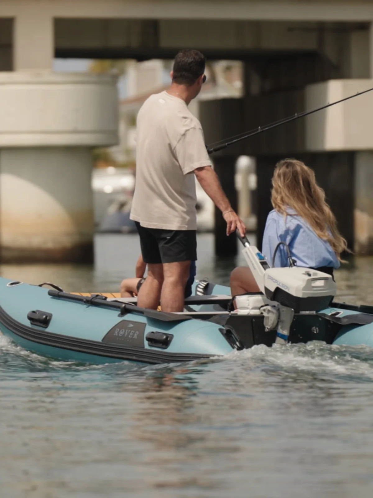 Two people on a small inflatable boat with a motor, under a bridge.