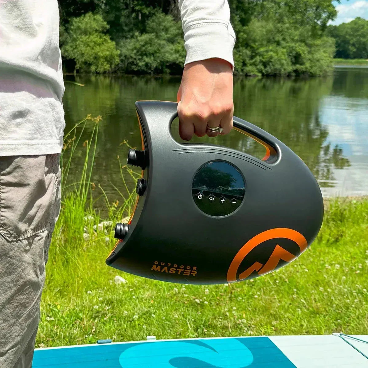 Person holding the OutdoorMaster Shark 2S electric pump by the handle near a lake, showing its portable design for inflating paddleboards and docks.