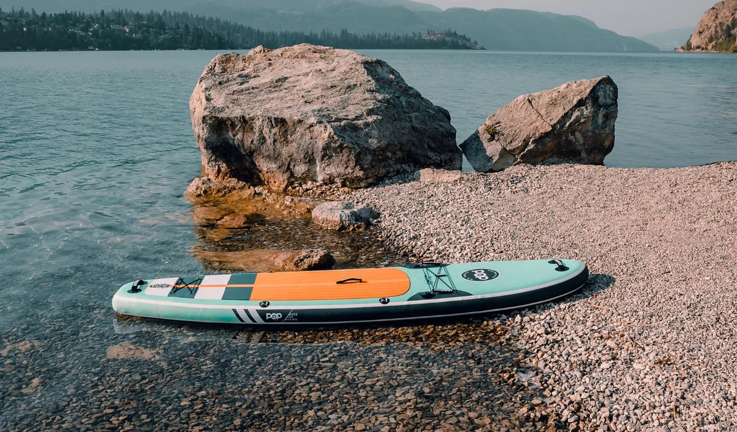POP Yacht Hopper 11'0 inflatable paddleboard in teak blue and mint resting by rocky shoreline.