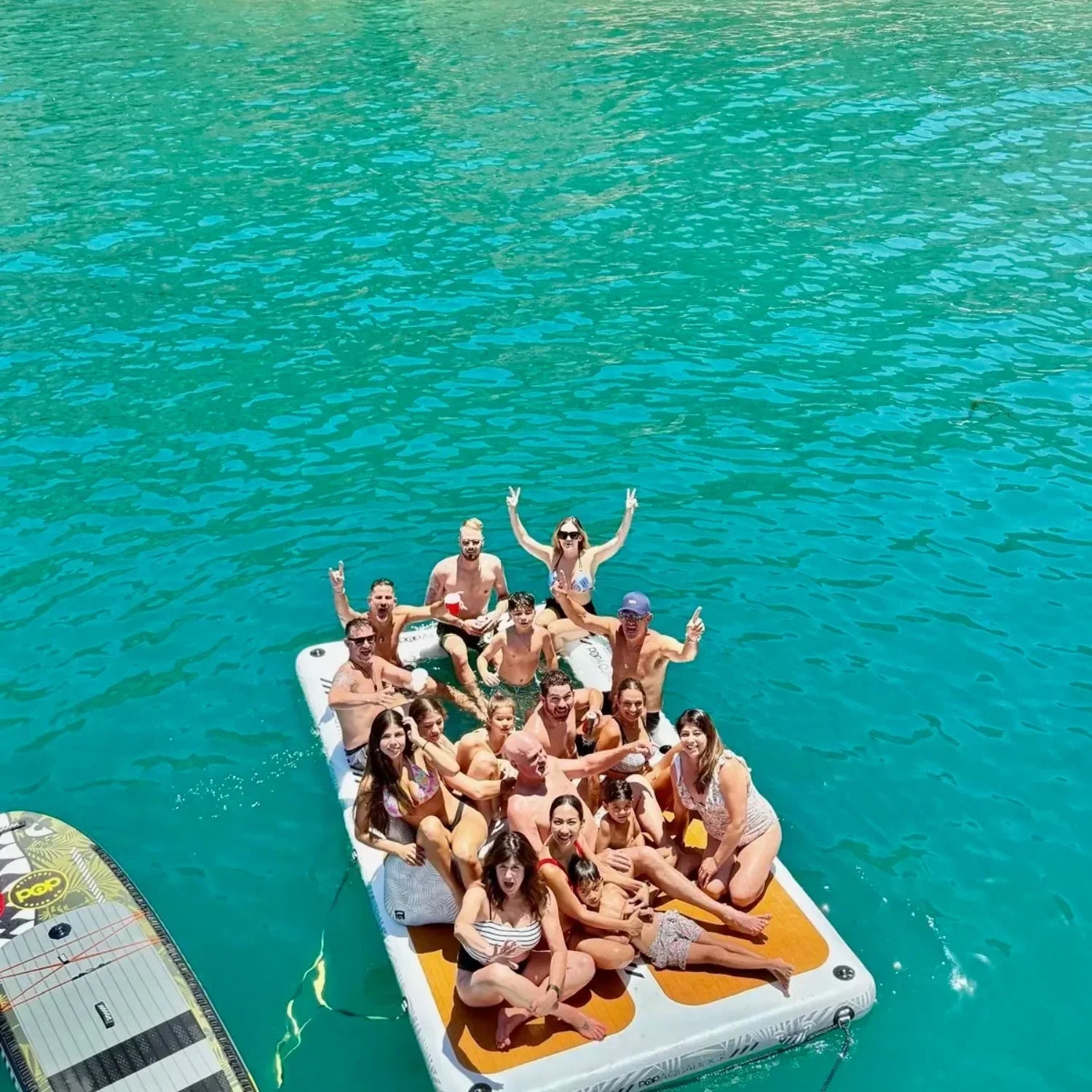 Large group of friends relaxing together on an Aqua Dock inflatable floating platform, enjoying the sun on clear turquoise water.