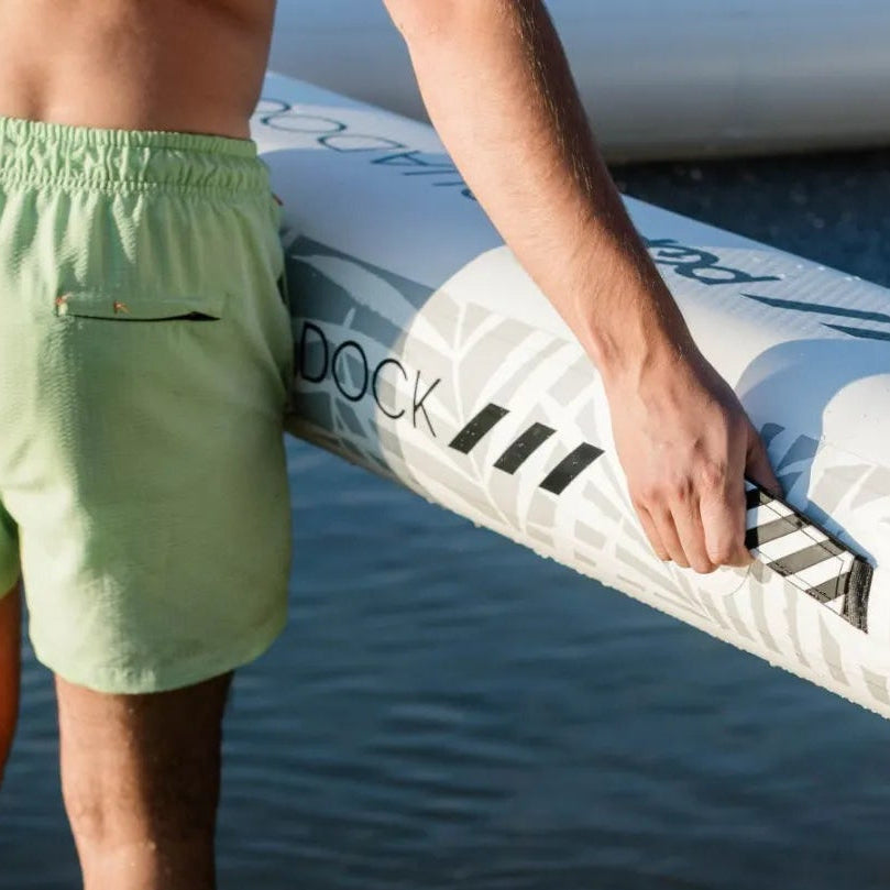 Close-up of a man carrying an Aqua Dock inflatable platform by its reinforced side handle while standing at the water’s edge.