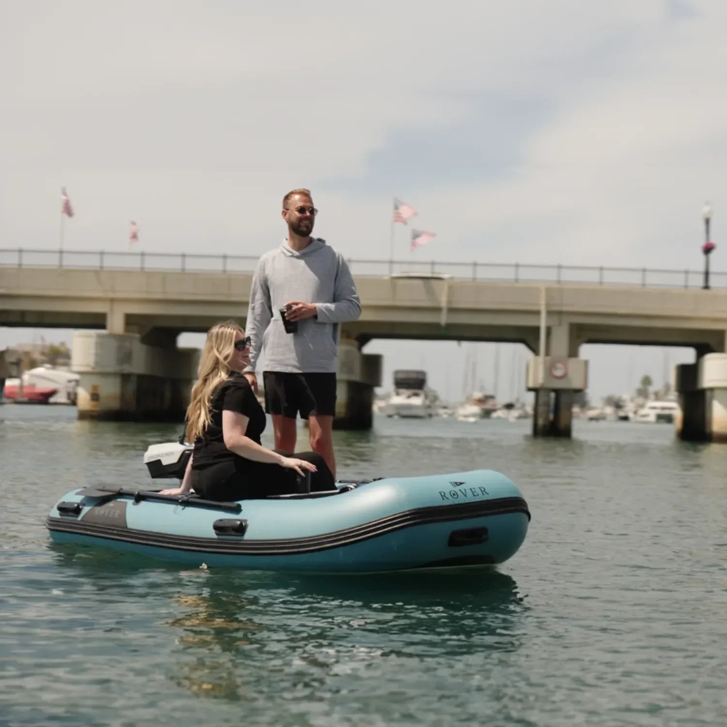 A couple cruising on a Rover Marine Battle Boat inflatable boat, passing under a bridge and a marina full of sailboats in the background.