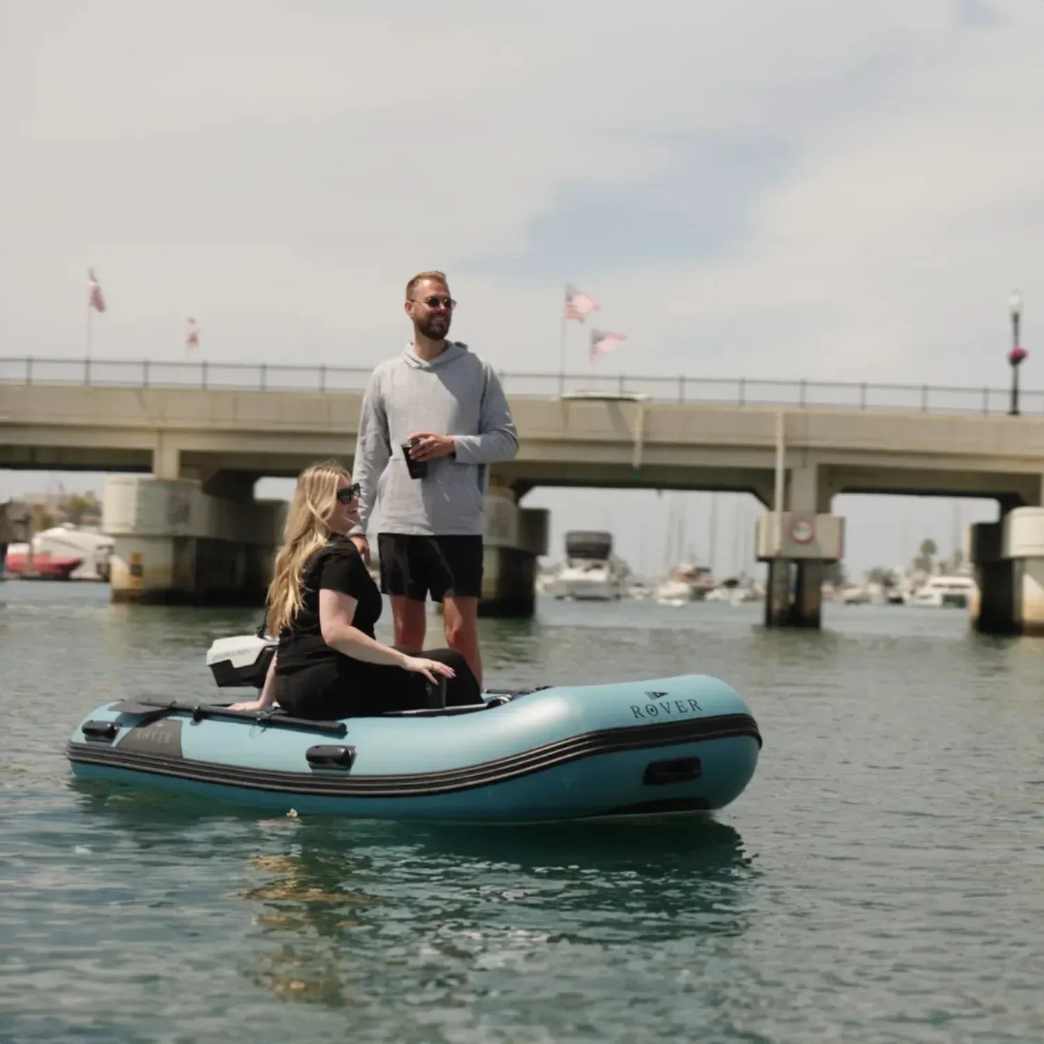 A couple cruising on a Rover Marine Battle Boat inflatable boat, passing under a bridge and a marina full of sailboats in the background.