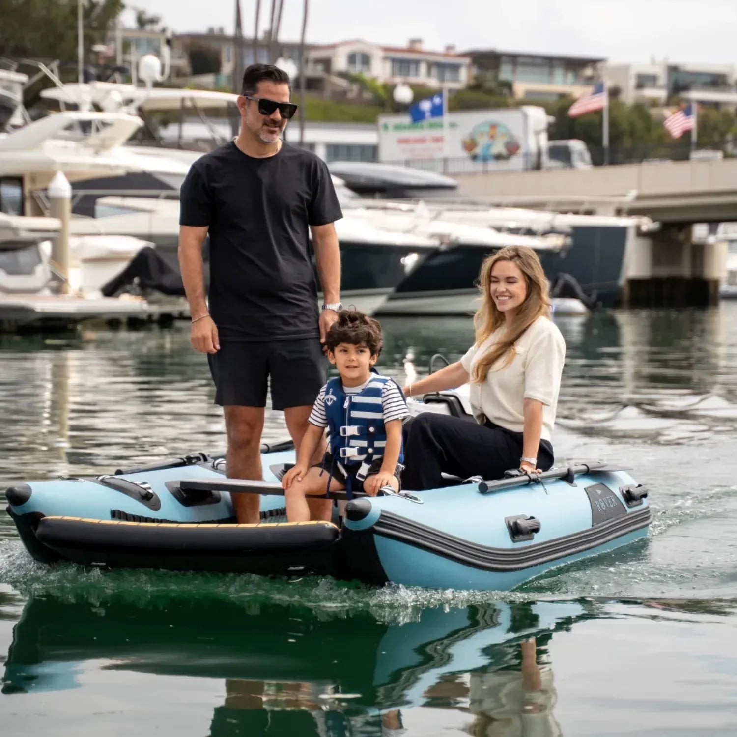 A family riding on a Rover Marine Battle Cat inflatable boat, with a man standing, a woman seated, and a child in a life jacket enjoying time together on the water.